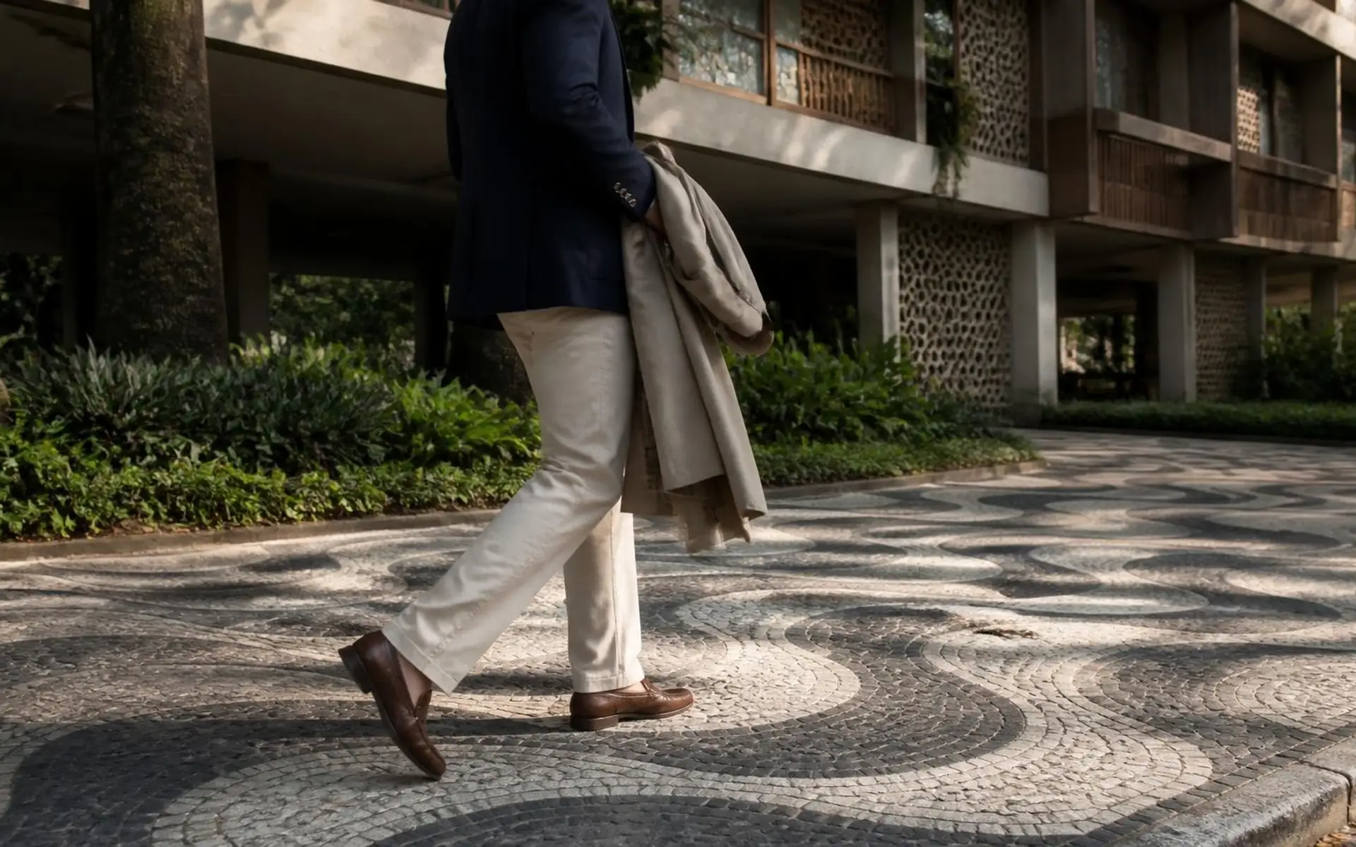 Profissional enquadrado do ombro para baixo caminha sobre calçada de pedra portuguesa em frente a edifício residencial modernista brasileiro com pilotis e cobogós